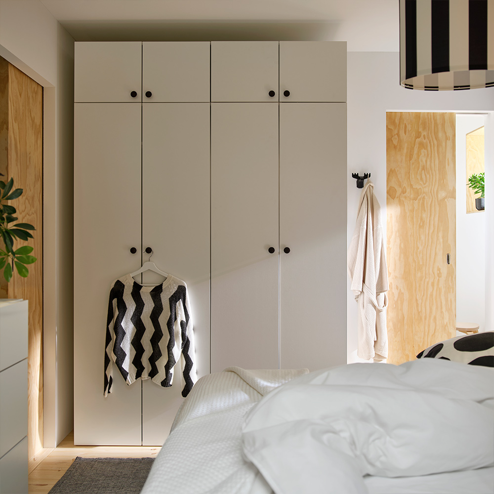 A sun-drenched bedroom with light-coloured walls and a rug, with LASTARE wardrobe combinations. A sweater hangs for the knob.