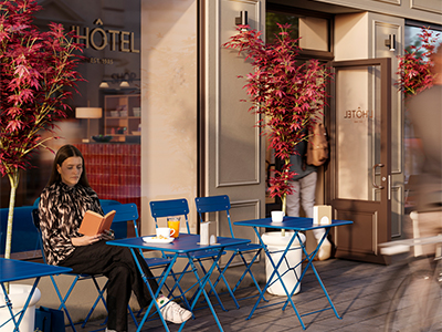 A woman sitting at a blue folding table outdoor cafe setup reading a book in front of a building named "L'HÔTEL", flanked by potted Japanese maple trees.