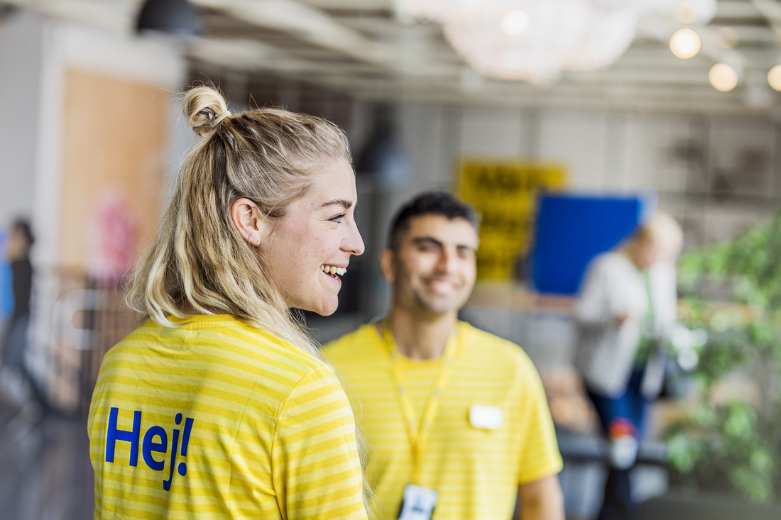 Close-up of a smiling IKEA coworker with blonde hair in a bun, wearing a yellow striped shirt with "Hej!" in blue. A blurred colleague stands in the background.