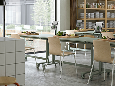 Long sage green tables on wheels set up with wooden chairs, cutting boards with ingredients, and digital screens, in a bright, modern workshop space.