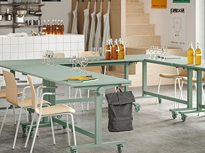 Sage green tables on wheels arranged in a U-shape with wooden chairs, wine glasses, and bottles, in a kitchen workshop or tasting room space.