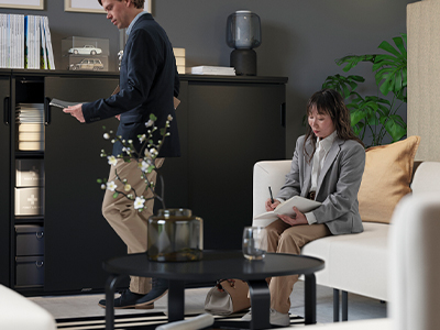A professional office setting where a woman sits on a beige sofa writing in a notebook, while a man in a blazer stands next to large black storage cabinets.