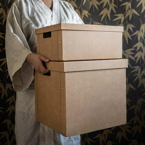 Persona con túnica blanca sosteniendo una pila de cajas de almacenamiento marrones con asas.