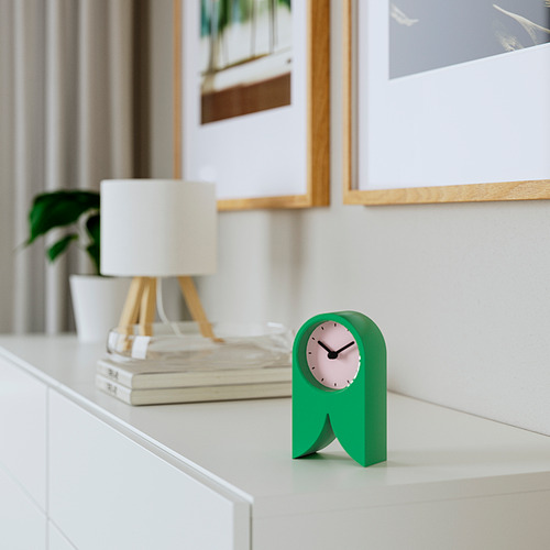 Un pequeño reloj FLODKRÄFTA verde está sobre el escritorio, silencioso, único. Cerca de la lámpara, libros, planta, arte. Ligero, de plástico.