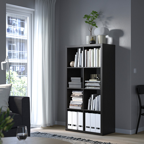 Estantería KALLAX negra con libros, decoración y carpetas junto a la ventana. Silla, cortina, copa de vino cerca.
