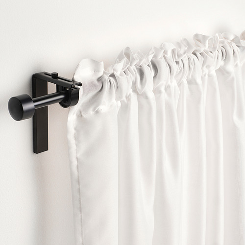 La barra de cortina negra sostiene cortinas blancas transparentes con cinta de encabezado para pliegues fáciles, montadas en una pared de color claro.