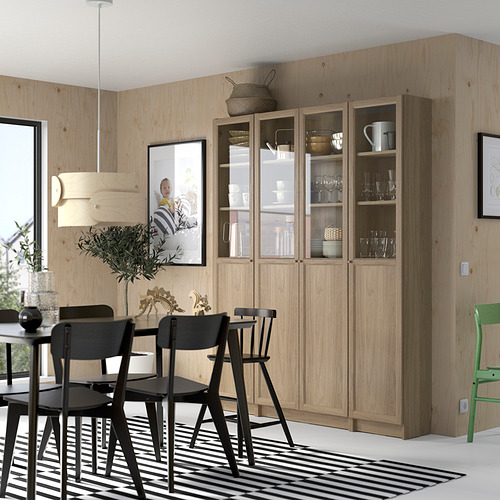 Comedor moderno con una gran estantería de madera BILLY, sillas negras, alfombra a rayas. Madera clara, puertas de vidrio para exhibir platos. Diseño simple y limpio.