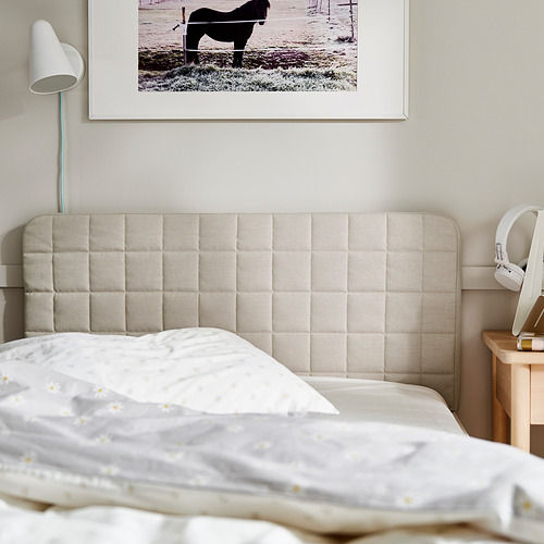 Cama de color neutro con cabecero acolchado, con una foto enmarcada encima. Una lámpara y unos auriculares descansan sobre una mesita de noche de madera.
