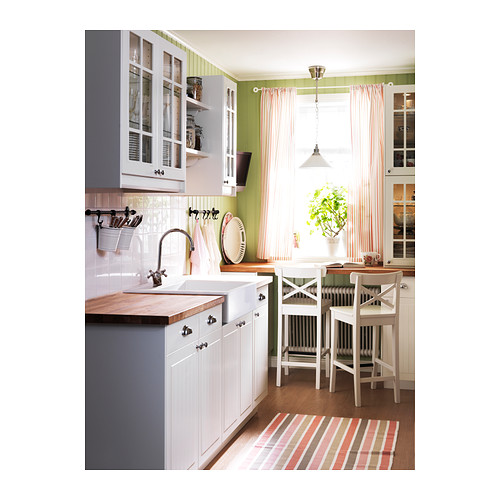Cocina luminosa con gabinetes blancos y verdes, cortinas a rayas, encimeras de madera y dos taburetes blancos junto a la ventana.