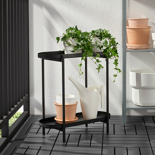 Soporte para plantas de metal negro con macetas blancas y plantas verdes exuberantes sobre suelo de baldosas grises, perfecto para uso en interiores y exteriores.