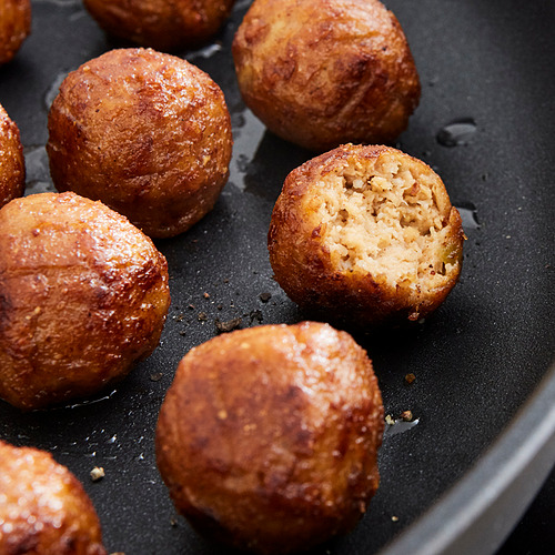 Patrones de buñuelos dorados, redondos y crujientes chisporroteando en una sartén negra. Uno está abierto, revelando un interior suave y parecido a la carne.