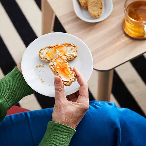 Persona sosteniendo tostadas con mermelada sobre mesa de madera. Suéter verde, camisa azul. Tarro de mermelada visible.