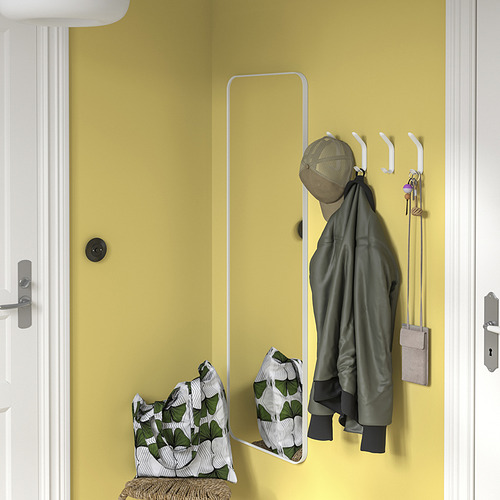 Pared amarilla con ganchos SNÖRAKA blancos que sostienen la chaqueta verde, la bolsa y las llaves. Espejo debajo, banco de mimbre con almohada.