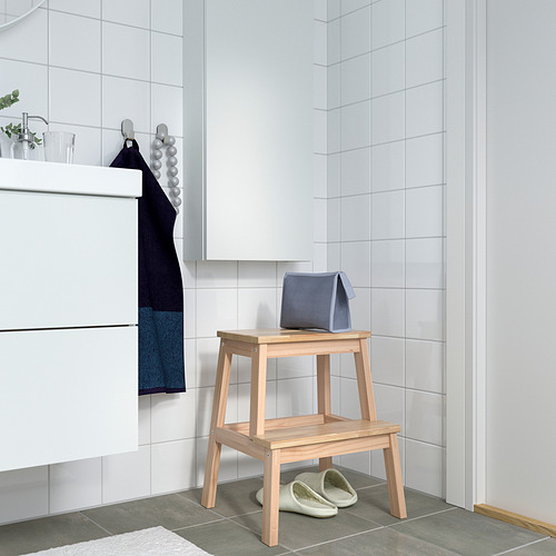 Baño minimalista con paredes de azulejos blancos. El taburete de madera BEKVÄM contiene toalla azul y zapatillas.