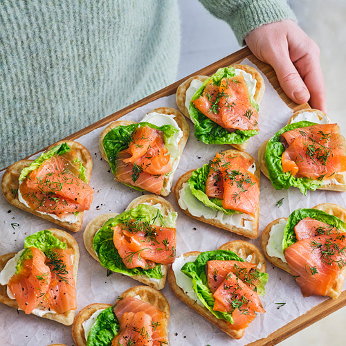 Aperitivos de waffles en forma de corazón cubiertos con salmón ahumado, lechuga y eneldo. Listo para hornear y servido en una tabla de madera.