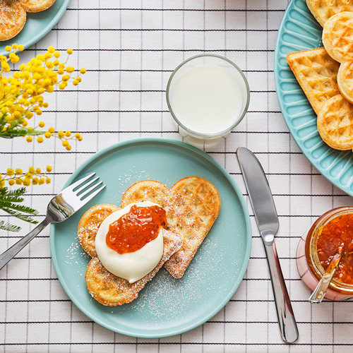 Gofres en forma de corazón con azúcar, nata, mermelada en plato turquesa con utensilios y flores.