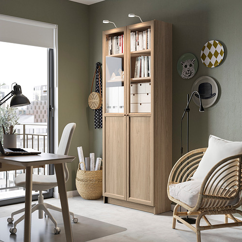 Acogedora oficina en casa con librería Billy, silla de mimbre, lámpara y jarrón. Luz natural, ambiente tranquilo.