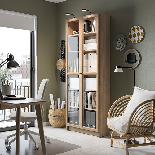 Librería BILLY de madera clara con puertas de vidrio, cerca de una silla blanca y una pequeña mesa, decorada con platos y jarrones.