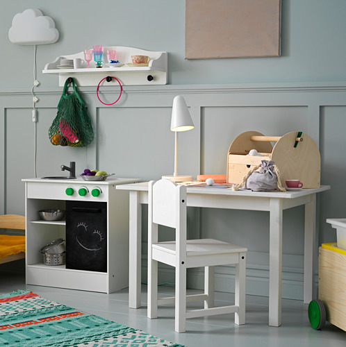 Sala de juegos con mesa de madera blanca, silla, cocina de juguete, lámpara y elementos decorativos.