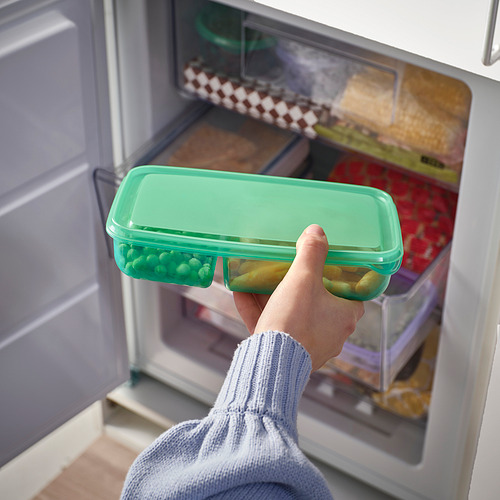 Sosteniendo de la mano un recipiente verde con alimentos, colocándolo en el refrigerador.