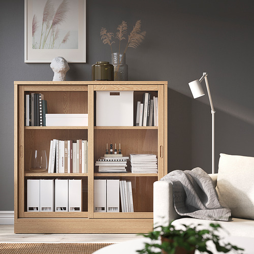 Acogedora sala de estar con librería de roble TONSTAD, llena de libros, y un sillón blanco, sobre paredes oscuras.