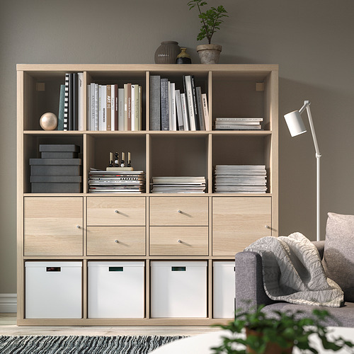Estantería de madera KALLAX con libros y decoración, cajones, sofá gris, lámpara blanca.