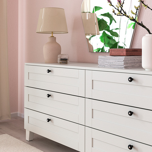 Cómoda blanca con seis cajones, pomos negros, lámpara y libros.