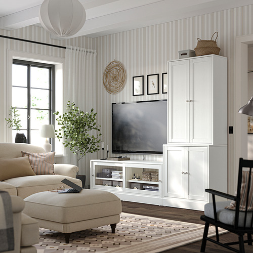 Sala de estar moderna con unidades de almacenamiento HAVSTA blancas, sofá beige, alfombra estampada y papel tapiz a rayas; TV y artículos decorativos expuestos.