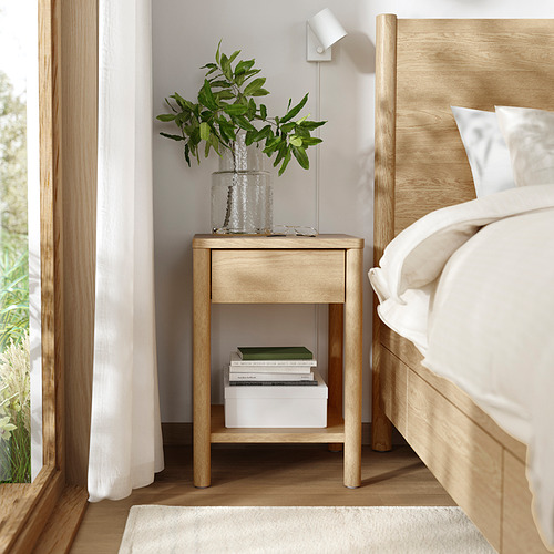 Mesita de noche de madera clara con cajón, libros y una planta.