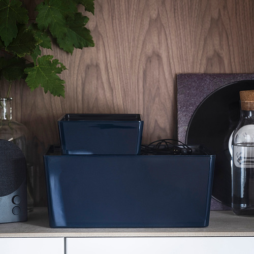 Dos cajas azules de almacenamiento en una estantería, una con tapa, junto a un altavoz y una botella.