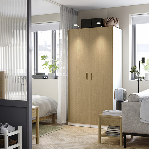 Un gran armario PAX de madera clara con bisagras de cierre suave junto a una cama, iluminado por la luz natural de una ventana cercana.