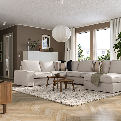 Sala de estar moderna con un gran sofá seccional beige KIVIK en ángulo contra la pared marrón, mesa de centro de madera y grandes ventanales.