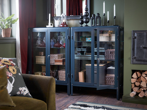 Sala de estar con mueble de metal y vidrio FABRIKÖR azul que muestra elementos de decoración, cerca de un sofá marrón con almohada floral.