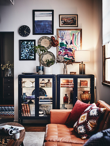 Sala de estar moderna con sofá de cuero naranja, almohadas estampadas, gabinete de vidrio negro con jarrones coloridos, arte y una lámpara.