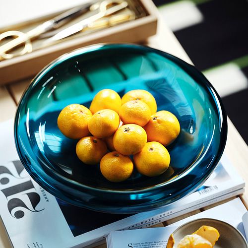 Un cuenco de vidrio azul lleno de pequeñas frutas redondas se encuentra encima de las revistas. La forma orgánica de los cuencos lo convierte en una pieza decorativa.