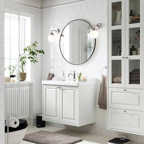 Baño blanco moderno: espejo redondo, lavabo de pared, vitrina, plantas y rollo de papel higiénico.