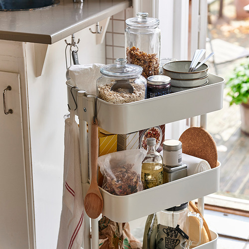 Carro de cocina con frascos de vidrio que almacenan alimentos, tazones y utensilios, colocado cerca de una ventana para facilitar el acceso y la exhibición.