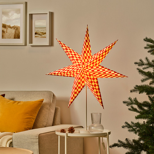 Linterna de estrella roja y amarilla STRÅLA cerca del árbol de Navidad, más alta que el sillón, sobre mesa blanca con piñas.