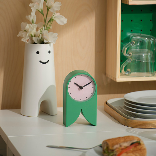 Reloj verde FLODKRÄFTA sobre encimera blanca con jarrón y sándwich.