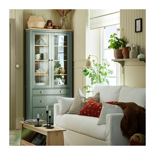 Sala de estar con sofá blanco, almohadas de colores, gabinete de madera en verde, estantes con plantas y decoraciones, ventana con cortina de luz.