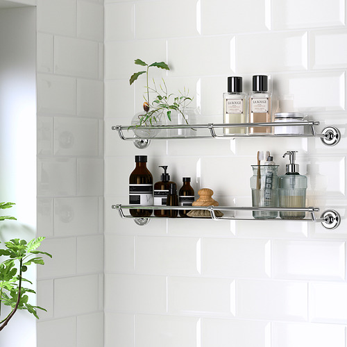 Los estantes de pared de metal con detalles cromados sostienen artículos de tocador y una planta, montados en una pared de azulejos blancos.