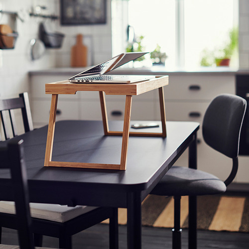 Mesa de madera para portátil sobre una mesa negra en una habitación luminosa, con una silla al lado.