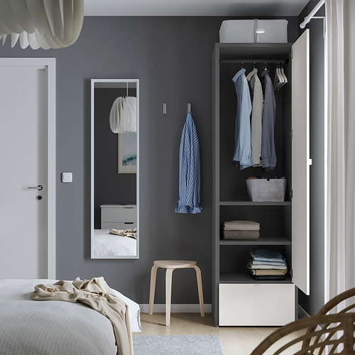 Dormitorio con armario abierto que muestra ropa, artículos doblados y un taburete. Cama blanca con manta beige en primer plano, y espejo en pared azul.