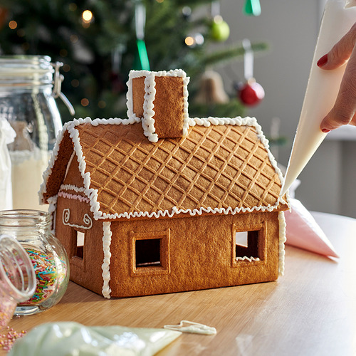 Una casa de jengibre decorada con glaseado blanco. La casa tiene un color marrón, techo con textura de gofre y una chimenea con líneas decorativas blancas.