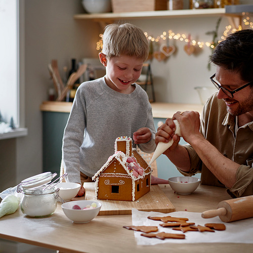 Hombre y niño decoran la casa de jengibre con glaseado, rodeados de dulces y cuencos en una mesa de madera.