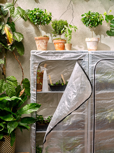 Pequeño invernadero con cubierta HYLLIS, que alberga varias plantas en macetas. Cubierta transparente, marco de metal, exuberante vegetación.