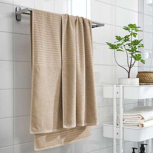 La toalla beige VÅGSJÖN cuelga en un estante, junto a estantes blancos con toallas dobladas y una planta.