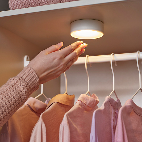 Una mano presionando la luz de un armario inalámbrico, iluminando camisas en perchas debajo de un estante.