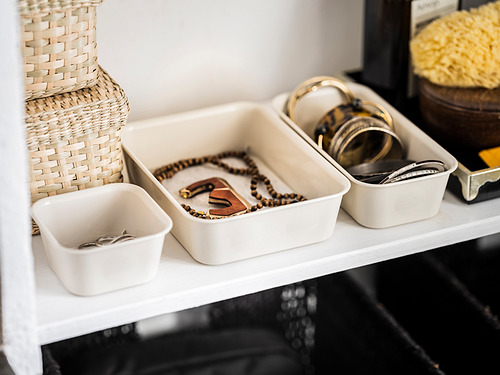 Estante blanco organizado con joyas en cajas de plástico transparente, con collares, anillos y pulseras.