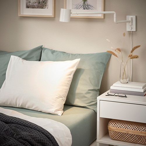 Rincón del dormitorio: sábana verde grisácea, almohadas blancas, manta gris. Mesa auxiliar con libros, plantas, lámpara de pared.